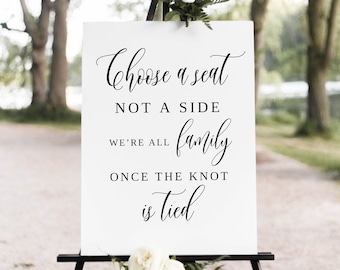 May include: White sign with black calligraphy text on an easel. The text reads "Choose a seat not a side we're all family once the knot is tied".