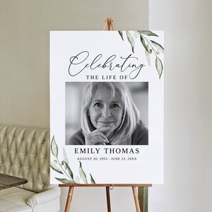 May include: A white memorial sign with green foliage border. The sign reads "Celebrating the Life of Emily Thomas August 20, 1953 - June 23, 20XX". A black and white photo of Emily is in the center of the sign.