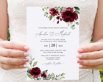 May include: A white wedding invitation with burgundy floral accents and elegant script. The invitation reads "Amelia & Michael" and includes the wedding date, time, and location. The invitation is held by a person wearing a white lace dress.