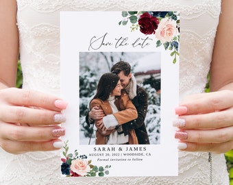 May include: Save the date card featuring a photograph of a couple in a snowy scene. The card is decorated with a floral design in shades of burgundy, pink, and blue. The text on the card reads "Save the date", "Sarah & James", "August 20, 2022 | Woodside, CA", and "Formal invitation to follow".