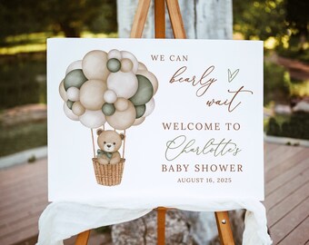 May include: A white sign with a brown teddy bear in a hot air balloon made of green and beige balloons. The sign reads "We can barely wait. Welcome to Charlotte's Baby Shower. August 16, 2025."