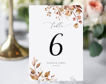May include: Table number six with a watercolor floral design in shades of brown and beige. The text "Table" is written in cursive above the number. The text "Sarah & James" and the date "10.14.2022" are printed below the number.