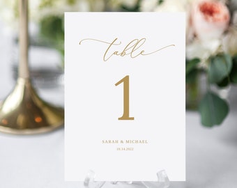 Könnte beinhalten: Weiße Tischnummerkarte mit goldener Kalligraphie, die "Tisch" und die Nummer "1" in Gold sagt. Die Karte enthält auch den Text "SARAH & MICHAEL" und "10.14.2022" in Schwarz.