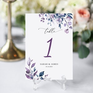 May include: Purple and blue watercolor floral table number card with the number "1" and the text "Table 1" in a cursive font. The text "SARAH & JAMES" and the date "10.14.2025" are printed below the number.