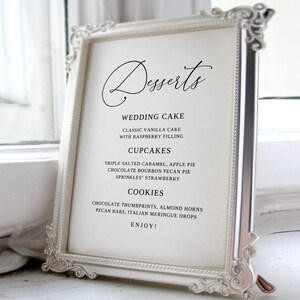 Könnte beinhalten: Ein weiß gerahmtes Schild mit schwarzem Text, das die Dessert-Optionen für einen Hochzeitsempfang auflistet. Das Schild lautet "Desserts", "Wedding Cake", "Classic Vanilla Cake with Raspberry Filling", "Cupcakes", "Triple Salted Caramel, Apple Pie, Chocolate Bourbon Pecan Pie, Sprinkles' Strawberry", "Cookies", "Chocolate Thumbprints, Almond Horns, Pecan Bars, Italian Meringue Drops", und "Enjoy!"