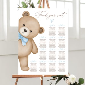 Può includere: Illustrazione di un orsacchiotto marrone chiaro con un papillon blu su un cartello bianco. Il cartello riporta le parole "Find your seat" e un elenco di nomi organizzati per numero di tavolo. Il cartello è esposto su un cavalletto di legno.