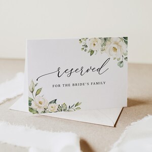 May include: A white card with the word "reserved" in elegant script, with the text "FOR THE BRIDE'S FAMILY" below. The card is decorated with watercolor floral accents of white roses and green leaves. It rests on a white envelope.