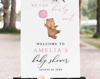 Op de afbeelding: Een wit babyborrel welkomstbord met de tekst "We can bearly wait" en "Welcome to Amelia's baby shower". Een bruine teddybeer houdt een roze ballon vast. Het bord staat op een zwart schildersezel met witte bloemen.
