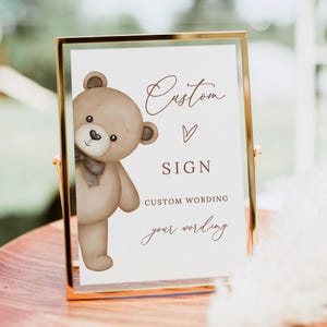 Könnte beinhalten: Ein goldgerahmtes Schild mit einem Teddybär-Motiv und dem Text "Custom Sign". Der Bär ist hellbraun mit einer grauen Fliege. Das Schild bietet Platz für individuelle Texte. Ideal für Babypartys oder Kinderzimmer.