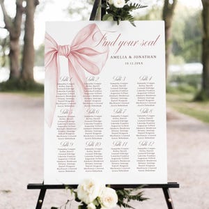 Könnte beinhalten: Ein Hochzeits-Sitzplan auf einer Staffelei. Das Schild zeigt eine rosa Schleife und die Worte "Find your seat". Namen sind unter den Tischnummern 1-12 aufgeführt. Das Hochzeitsdatum wird ebenfalls angezeigt.