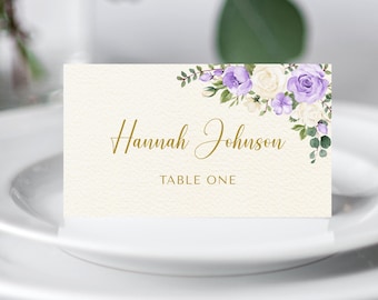 Puede incluir: Una tarjeta de mesa color crema con el nombre "Hannah Johnson" en escritura dorada y "TABLE ONE" debajo. La esquina superior derecha presenta un arreglo floral de flores moradas y blancas con hojas verdes. La tarjeta está sobre un plato blanco.