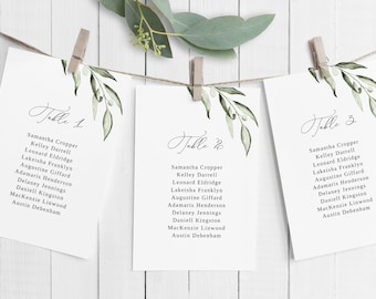 May include: Three white cards with green foliage illustrations and handwritten calligraphy. The cards are labeled "Table 1", "Table 2", and "Table 3" and list names for each table.