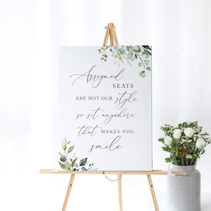 May include: A white sign with green foliage on a wooden easel. The sign reads "Assigned Seats Are Not Our Style So Sit Anywhere That Makes You Smile".