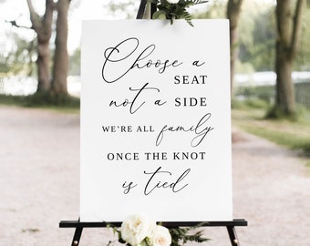 May include: White sign with black calligraphy text on an easel. The text reads "Choose a seat not a side. We're all family once the knot is tied."