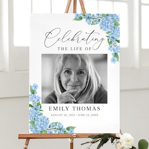 May include: A white memorial sign with a black and white photo of a woman. The sign has blue hydrangea flowers on the sides and the text "Celebrating the life of Emily Thomas August 20, 1953 - June 23, 20XX".