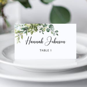 May include: A white place card with the name "Hannah Johnson" in elegant black script, accented with watercolor greenery. The card also displays "TABLE 1". The card is set on a white plate.