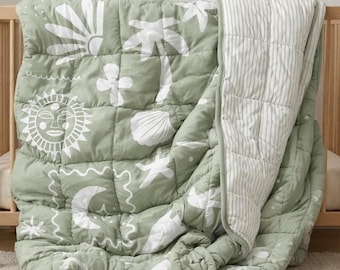 May include: A sage green quilt with white tropical-themed designs, including a sun, palm trees, and seashells. The quilt is quilted in a grid pattern and has a striped reverse side. The quilt is draped over a wooden crib.