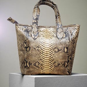 Puede incluir: Un bolso tote con estampado de piel de serpiente con dos asas y un tirador de cremallera. El bolso es principalmente de color beige con marcas marrón oscuro y negro. El bolso está sobre una superficie blanca.