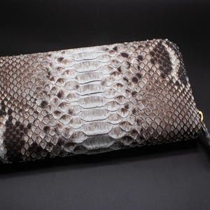 Peut inclure: Un portefeuille rectangulaire avec un motif peau de serpent dans les tons de gris, blanc et noir. Le portefeuille a une fermeture éclair avec une tirette dorée et une petite dragonne. Le design général est élégant et texturé.