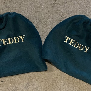 Peut inclure: Deux sacs &agrave; cordon bleu sarcelle avec le mot "TEDDY" brod&eacute; en or sur le devant.