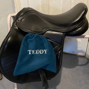 Peut inclure: Une selle en cuir noir avec un tapis de selle bleu sur lequel est brod&eacute; "TEDDY" en blanc.
