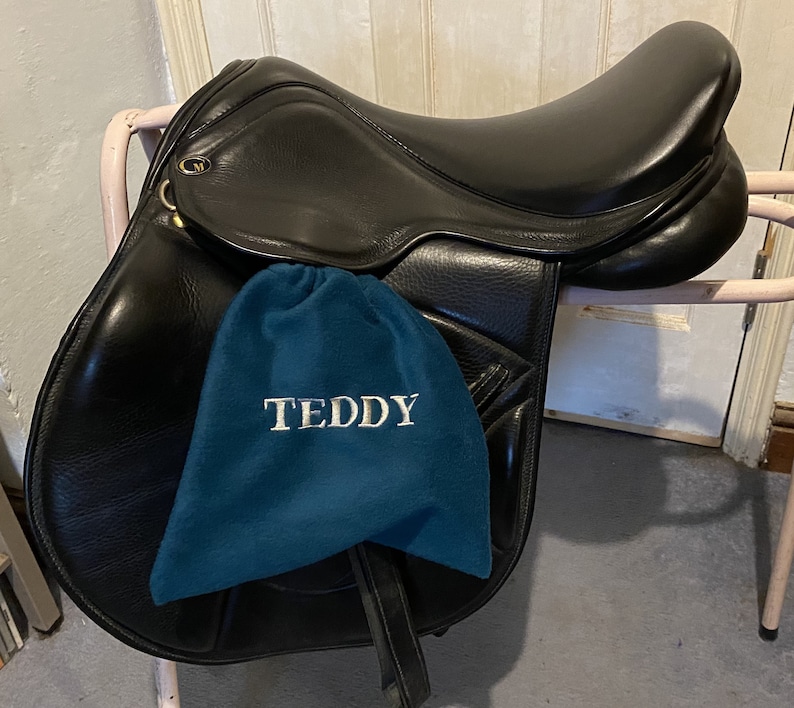Peut inclure: Une selle en cuir noir avec un tapis de selle bleu sur lequel est brod&eacute; "TEDDY" en blanc.