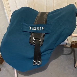 Peut inclure: Selle de cheval en tissu bleu sarcelle avec des sangles noires et une boucle argent&eacute;e. La selle est brod&eacute;e du nom "TEDDY" en blanc.