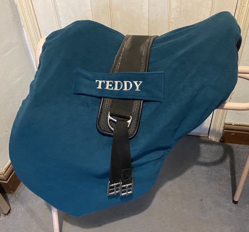 Peut inclure: Selle de cheval en tissu bleu sarcelle avec des sangles noires et une boucle argent&eacute;e. La selle est brod&eacute;e du nom "TEDDY" en blanc.