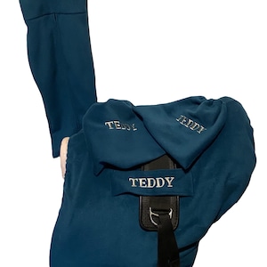 Puede incluir: Una funda de silla de montar azul turquesa con el nombre "Teddy" bordado. La funda tiene una solapa que cubre los estribos de la silla.