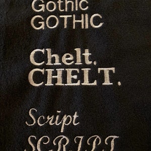 Peut inclure: Un &eacute;chantillon de tissu noir avec trois styles de police diff&eacute;rents brod&eacute;s en fil blanc. La premi&egrave;re police est &eacute;tiquet&eacute;e "Gothic", la deuxi&egrave;me est &eacute;tiquet&eacute;e "Chelt" et la troisi&egrave;me est &eacute;tiquet&eacute;e "Script".