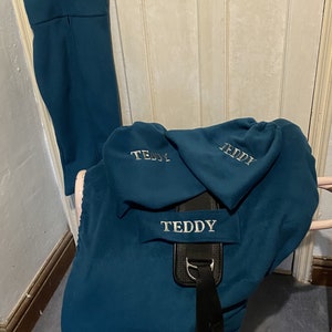 Peut inclure: Une housse de selle bleu sarcelle avec le nom "Teddy" brod&eacute; dessus. La housse est en polaire et poss&egrave;de une sangle noire avec des boucles. Il y a trois housses plus petites bleu sarcelle avec le nom "Teddy" brod&eacute; dessus.