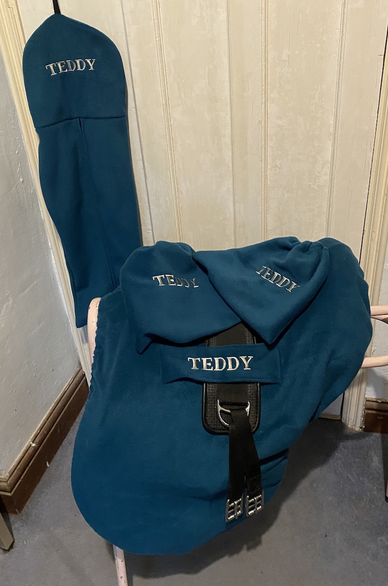Peut inclure: Une housse de selle bleu sarcelle avec le nom "Teddy" brod&eacute; dessus. La housse est en polaire et poss&egrave;de une sangle noire avec des boucles. Il y a trois housses plus petites bleu sarcelle avec le nom "Teddy" brod&eacute; dessus.