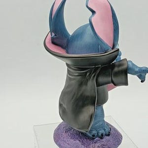 May include: A blue figurine of a cartoon character with large ears, wearing a black cloak. The ears are pink inside. The figurine is standing on a purple base.