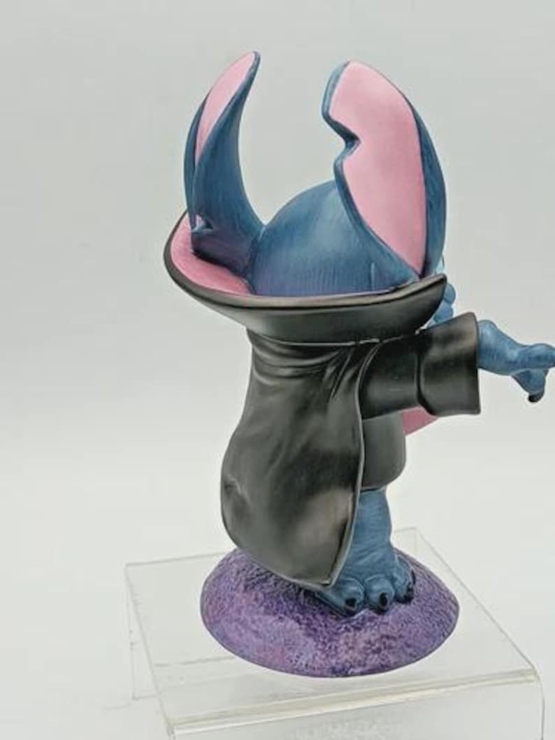 May include: A blue figurine of a cartoon character with large ears, wearing a black cloak. The ears are pink inside. The figurine is standing on a purple base.