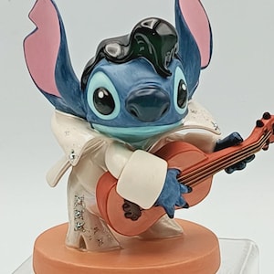 K&ouml;nnte beinhalten: Eine blaue Stitch-Figur, die als Elvis Presley verkleidet ist und eine rote Akustikgitarre spielt. Die Figur hat gro&szlig;e Ohren mit rosa Innenseiten, einen wei&szlig;en Overall und eine schwarze Pompadour-Frisur. Der Sockel ist hellorange.