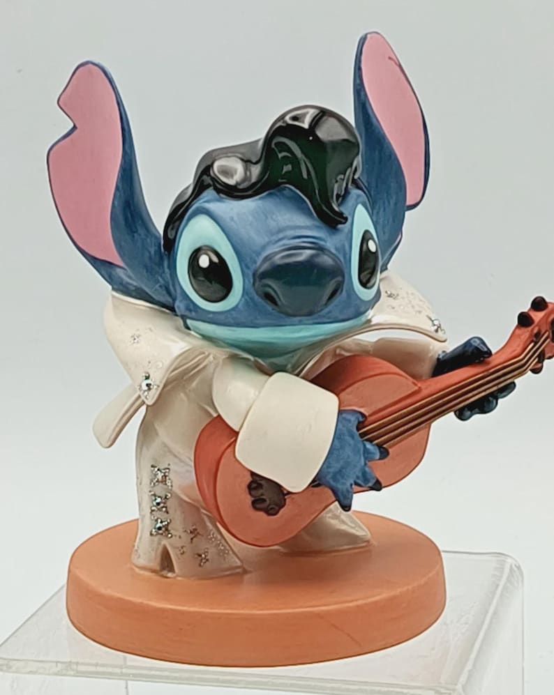 K&ouml;nnte beinhalten: Eine blaue Stitch-Figur, die als Elvis Presley verkleidet ist und eine rote Akustikgitarre spielt. Die Figur hat gro&szlig;e Ohren mit rosa Innenseiten, einen wei&szlig;en Overall und eine schwarze Pompadour-Frisur. Der Sockel ist hellorange.