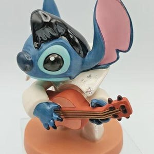 K&ouml;nnte beinhalten: Eine blau-schwarze Figur von Stitch, die Ukulele spielt. Die Figur tr&auml;gt ein wei&szlig;es Hemd mit Kragen und steht auf einem runden, orangefarbenen Sockel. Die Ohren sind rosa und blau.