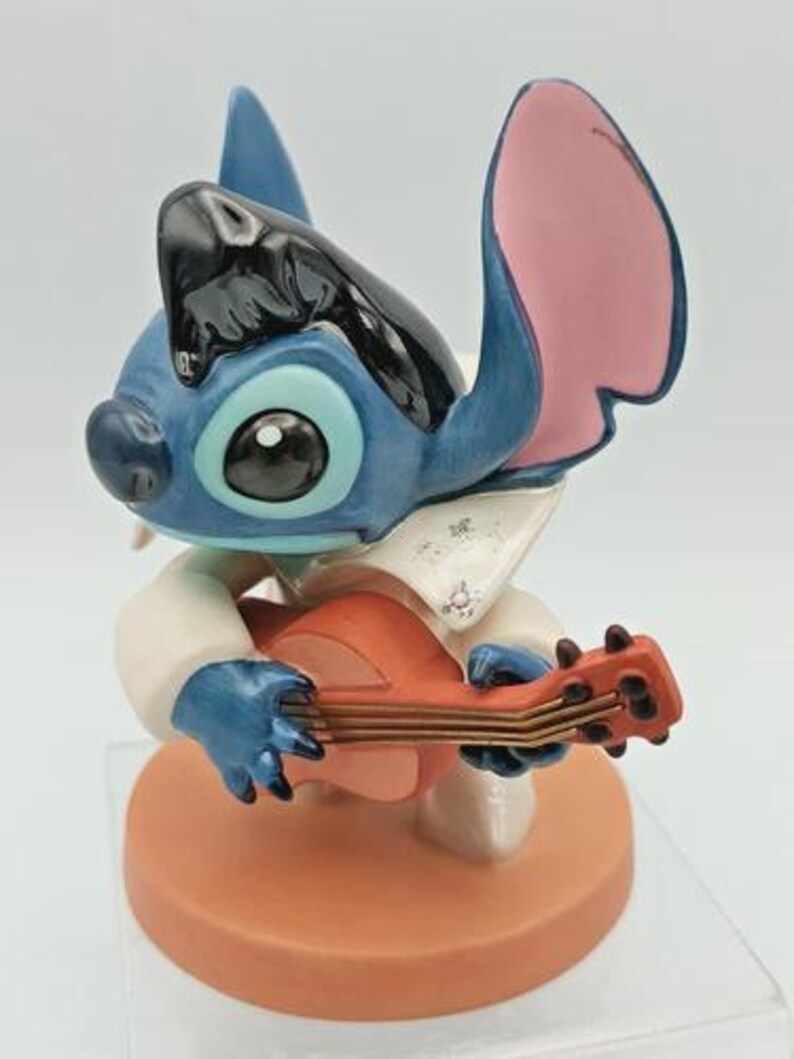 K&ouml;nnte beinhalten: Eine blau-schwarze Figur von Stitch, die Ukulele spielt. Die Figur tr&auml;gt ein wei&szlig;es Hemd mit Kragen und steht auf einem runden, orangefarbenen Sockel. Die Ohren sind rosa und blau.