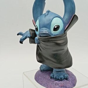 May include: A blue figurine of the character Stitch, wearing a black cape. The figurine has large ears, big eyes, and is standing on a purple base. The figure is posed with one arm outstretched.