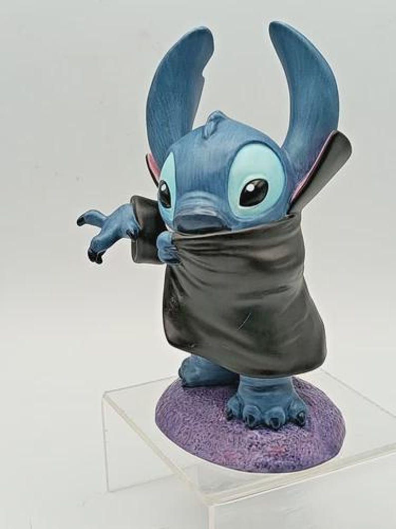 May include: A blue figurine of the character Stitch, wearing a black cape. The figurine has large ears, big eyes, and is standing on a purple base. The figure is posed with one arm outstretched.