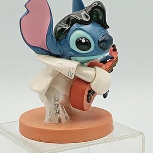K&ouml;nnte beinhalten: Blaue Stitch-Figur, bekleidet mit einem wei&szlig;en Overall mit silbernen Verzierungen, h&auml;lt eine rote Gitarre. Stitch hat schwarzes Haar und steht auf einer orangefarbenen, runden Basis. Ein Sammlerst&uuml;ck.