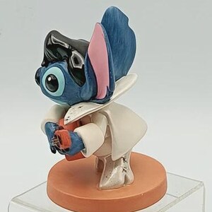 K&ouml;nnte beinhalten: Blaue und schwarze Figur einer Zeichentrickfigur mit gro&szlig;en Ohren und wei&szlig;em Outfit. Die Figur h&auml;lt ein kleines rotes Objekt und steht auf einem runden, orangefarbenen Sockel.