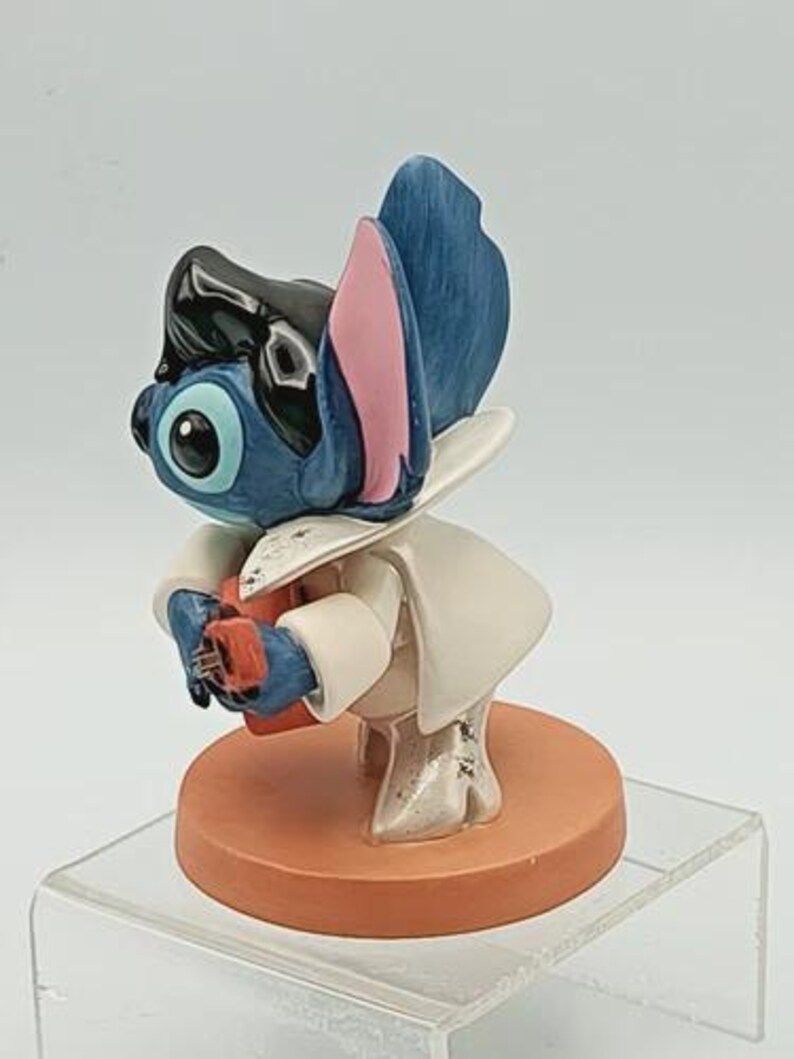 K&ouml;nnte beinhalten: Blaue und schwarze Figur einer Zeichentrickfigur mit gro&szlig;en Ohren und wei&szlig;em Outfit. Die Figur h&auml;lt ein kleines rotes Objekt und steht auf einem runden, orangefarbenen Sockel.