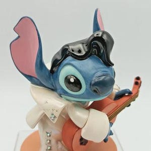 K&ouml;nnte beinhalten: Blaue Stitch-Figur, im Stil von Elvis Presley, spielt eine rote Ukulele. Die Figur hat schwarzes Haar, einen wei&szlig;en Overall mit glitzernden Akzenten und einen pfirsichfarbenen Sockel. Die Ohren sind rosa und blau.