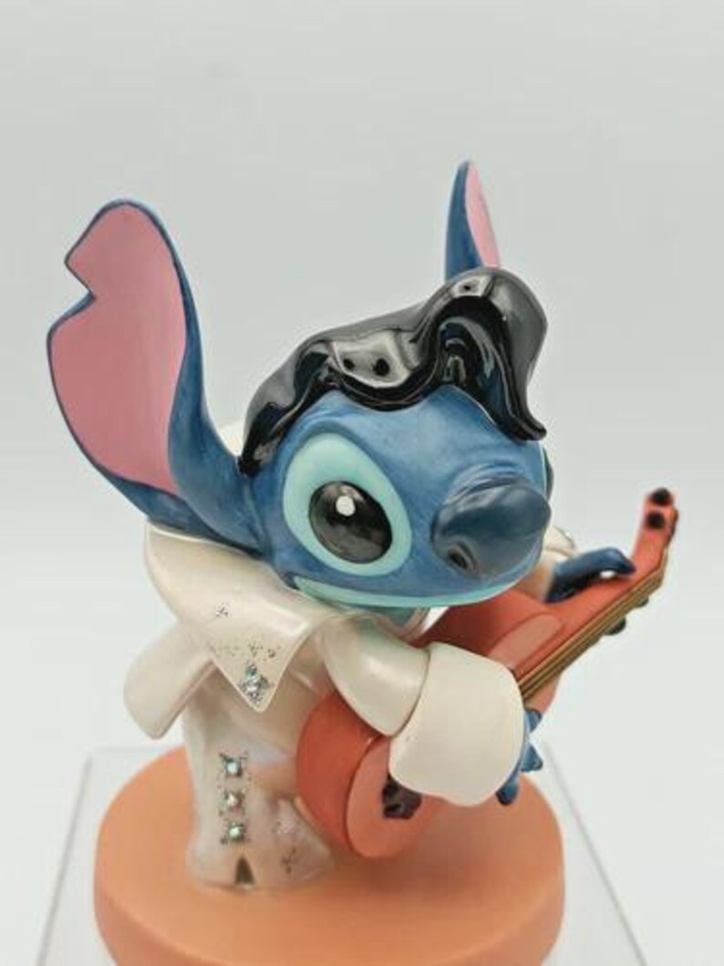 K&ouml;nnte beinhalten: Blaue Stitch-Figur, im Stil von Elvis Presley, spielt eine rote Ukulele. Die Figur hat schwarzes Haar, einen wei&szlig;en Overall mit glitzernden Akzenten und einen pfirsichfarbenen Sockel. Die Ohren sind rosa und blau.