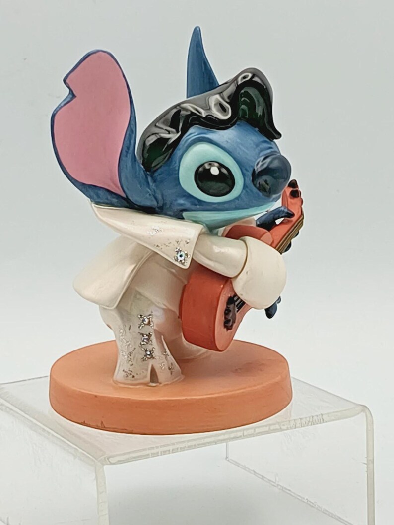 K&ouml;nnte beinhalten: Blaue Stitch-Figur, bekleidet mit einem wei&szlig;en Overall mit Strasssteinen und einer schwarzen Per&uuml;cke, spielt eine rote Gitarre. Die Figur hat gro&szlig;e schwarze Augen und rosa Ohren, auf einem orangefarbenen Sockel.