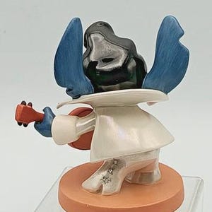 K&ouml;nnte beinhalten: Eine Figur von Stitch, einer Disney-Figur, die Ukulele spielt. Die Figur hat blaue Ohren, einen schwarzen Kopf und ein wei&szlig;es Outfit. Die Ukulele ist rot und braun, und die Basis ist orange.