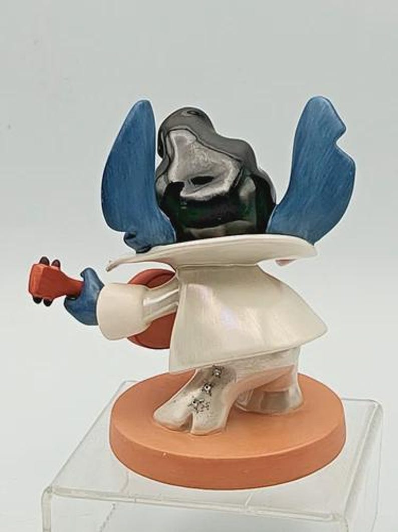 K&ouml;nnte beinhalten: Eine Figur von Stitch, einer Disney-Figur, die Ukulele spielt. Die Figur hat blaue Ohren, einen schwarzen Kopf und ein wei&szlig;es Outfit. Die Ukulele ist rot und braun, und die Basis ist orange.