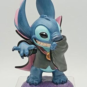 May include: A blue figurine of the character Stitch dressed as a vampire, with a black cape and pink accents. The figurine is standing on a purple base and has a mischievous grin.