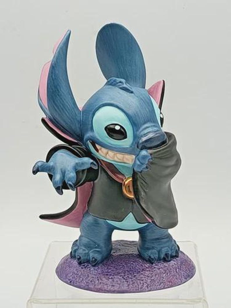 May include: A blue figurine of the character Stitch dressed as a vampire, with a black cape and pink accents. The figurine is standing on a purple base and has a mischievous grin.
